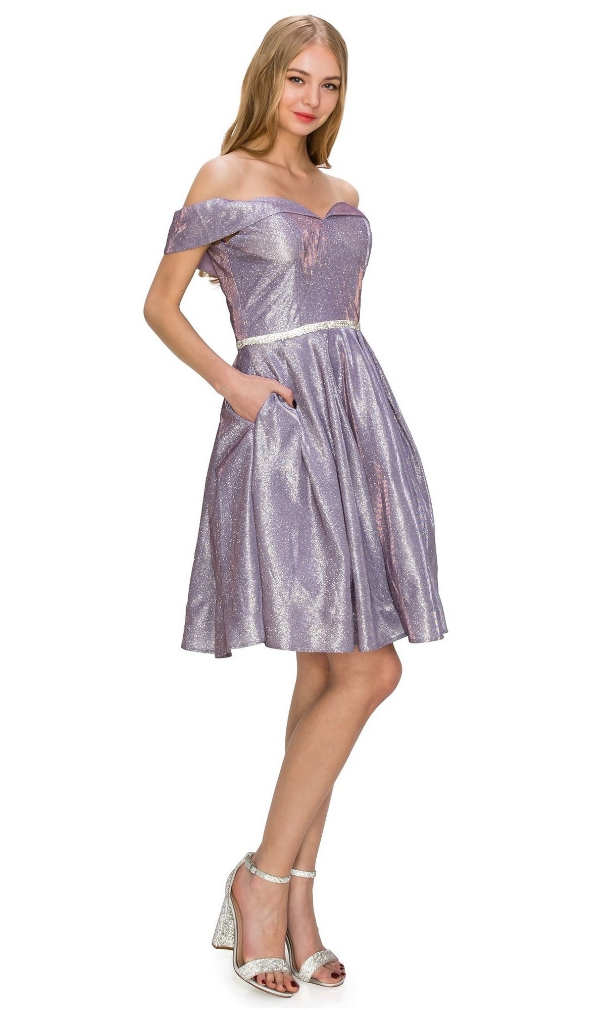 A person models the Off-Shoulder Short Glitter Homecoming Dress 8012J in lilac, featuring pockets, paired with silver high-heeled sandals, standing against a white background.